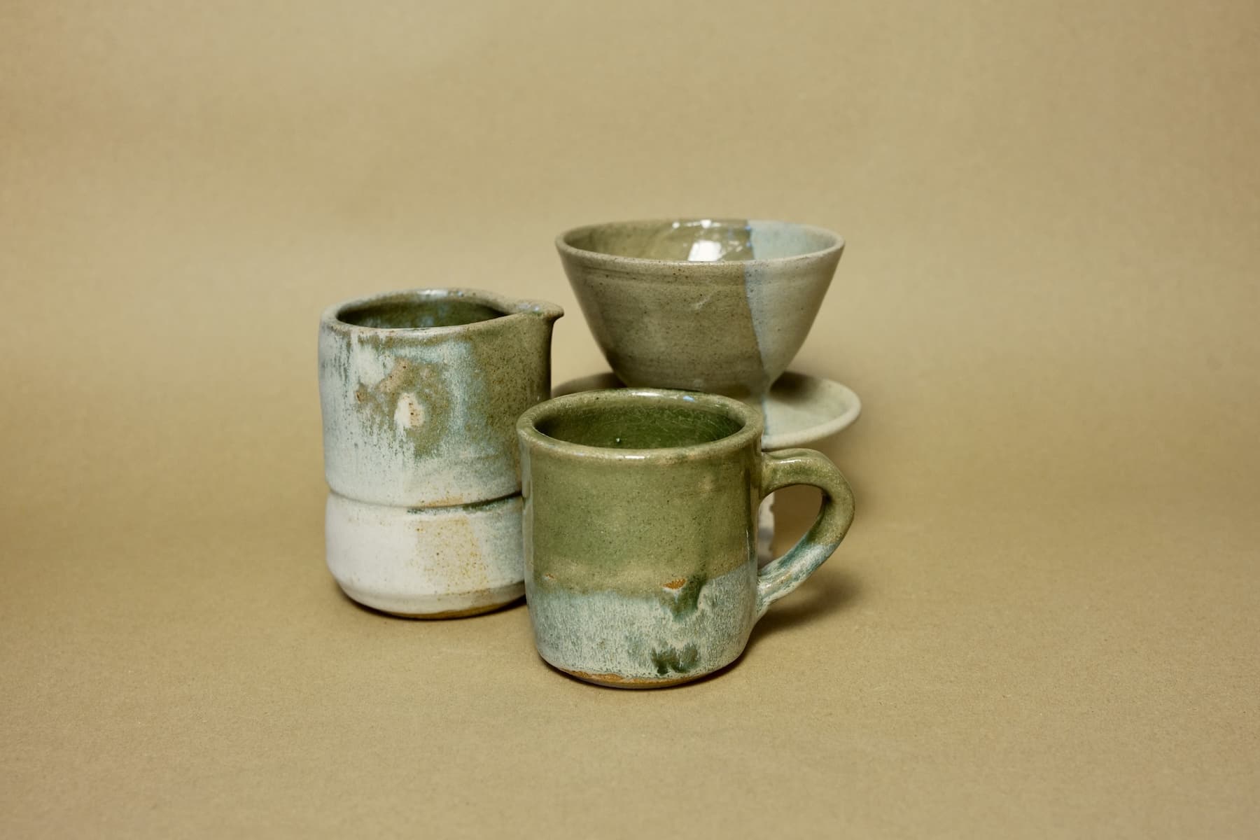 Close-up of a hand-thrown ceramic mug in sage green glaze, grouped with a matching coffee carafe and pour-over dripper.
